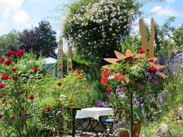 Ein Garten mit bunten Blumen und Pflanzen, der einen zentralen Tisch mit einer Decke und Stühlen zeigt, der vor Bäumen und einem bewölkten Himmel am Chelsea Flower Show steht.