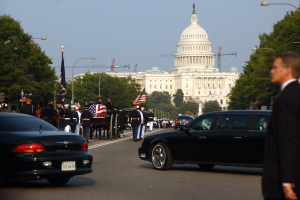 Ein Trauerzug mit Menschen in Mützen und mit Fahnen an der Capitol Building vorbeiziehend, begleitet von Fahrzeugen, Laternenpfählen, Bäumen und Gebäuden unter einem klaren Himmel.