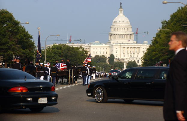 Ein Trauerzug mit Menschen in Mützen und mit Fahnen an der Capitol Building vorbeiziehend, begleitet von Fahrzeugen, Laternenpfählen, Bäumen und Gebäuden unter einem klaren Himmel.
