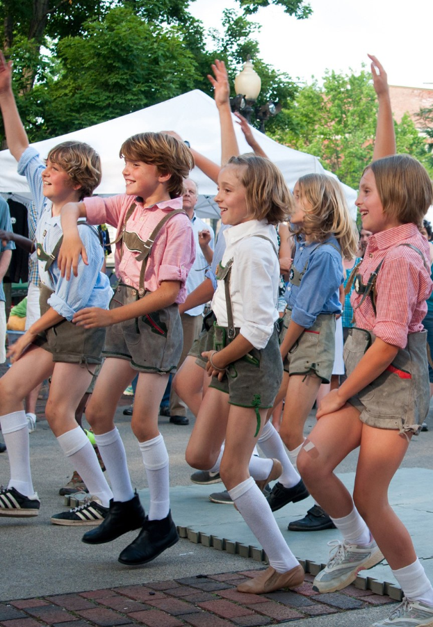 Kinder in traditioneller bayrischer Tracht, die auf einer Straße tanzen, umgeben von einer Menge, Zelten, Gebäuden und Bäumen unter einem klaren blauen Himmel.