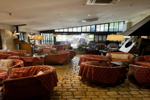 Ein geräumiger Empfangsbereich des Hotel de Ville mit zahlreichen Stühlen und Tischen, gepolsterten Sofas, Wandlampen, einer Deckenklimaanlage und großen Glasfenstern an der Rückseite.