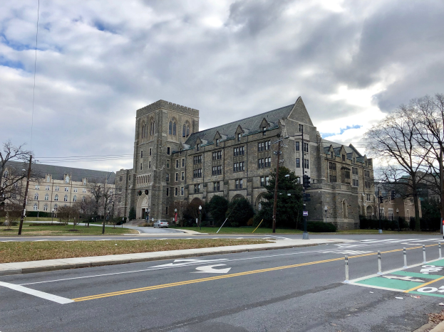 Großes Gebäude mit zahlreichen Fenstern, als der neue Campus der Universität Pennsylvania bezeichnet, umgeben von Bäumen, Straßenlaternen, Verkehrszeichen und Fahrzeugen unter einem bewölkten Himmel.