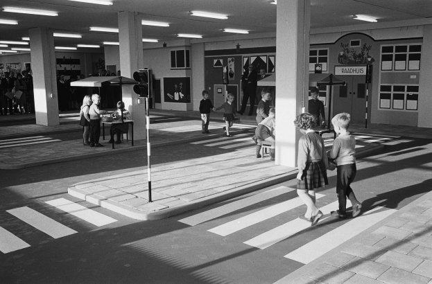 Schwarzes und weißes Foto von Kindern, die eine Straße an einer Ampel überqueren, mit Menschen auf Bänken, einer Verkehrsampel und Hintergrundsäulen mit Deckenleuchten.