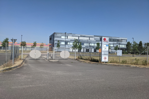 Großes industrielles Gebäude mit der Aufschrift "First Solar" und vielen Fenstern, umgeben von einem Metallzaun, Bäumen, Pflanzen, Gras, Schildern, Straßenlaternen und einem Weg unter einem bewölkten Himmel.