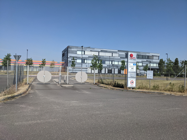 Großes industrielles Gebäude mit der Aufschrift "First Solar" und vielen Fenstern, umgeben von einem Metallzaun, Bäumen, Pflanzen, Gras, Schildern, Straßenlaternen und einem Weg unter einem bewölkten Himmel.