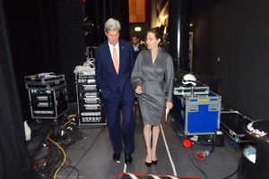 Angelina Jolie und John Kerry auf einem Boden umgeben von einer Gruppe von Menschen, verschiedenen Geräten, Kabeln, einem Helm auf einem Wagen, einem Müllcontainer, einem Vorhang, einer Säule und einer Decke mit Deckenleuchten.