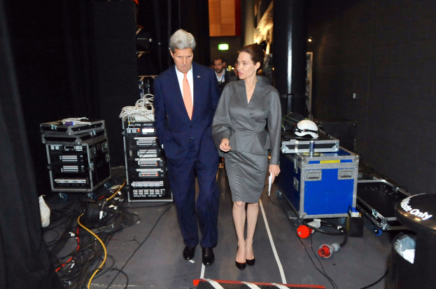 Angelina Jolie und John Kerry auf einem Boden umgeben von einer Gruppe von Menschen, verschiedenen Geräten, Kabeln, einem Helm auf einem Wagen, einem Müllcontainer, einem Vorhang, einer Säule und einer Decke mit Deckenleuchten.