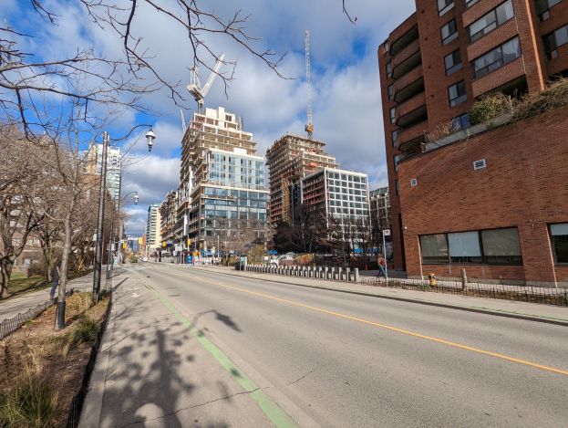 Eine Stadtstraße mit hohen Gebäuden, Straßenlaternen, Stromkabeln, Fahrzeugen, Fußgängern, Geländern, Bäumen und einem bewölkten Himmel.