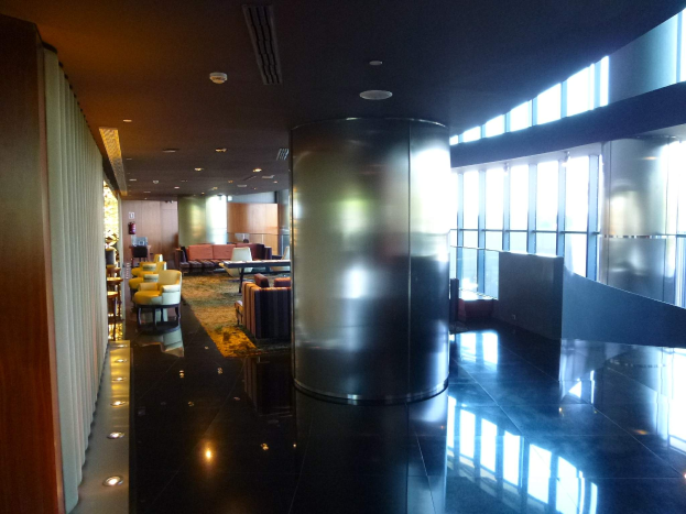 Eine geräumige First-Class-Lounge am Hong Kong International Airport mit großen Fenstern, hohen Decken mit elektrischem Licht, Sitzanordnungen einschließlich Stühlen und Sofas, Tischen, einem Teppichboden und einer zentralen Säule.