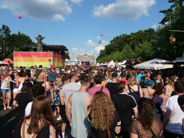Große Menschenmenge auf einer Straße mit Zelten, Bäumen, Pfählen, Lichtern und einer Statue während des Christopher Street Day in Berlin, mit Gebäuden, Wolken und Ballons im Hintergrund.