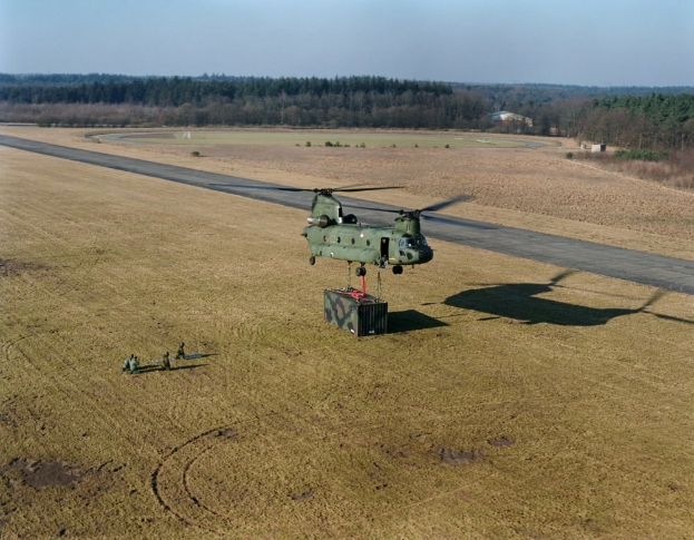 Militärhubschrauber fliegt über ein Feld neben einer Straße mit einigen Menschen unten, umgeben von Bäumen unter einem klaren blauen Himmel.