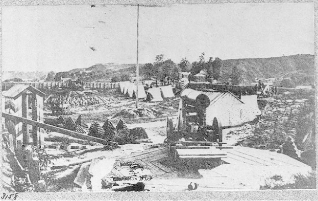 Schwarz-weißes Foto einer Baustelle mit einer Maschine im Vordergrund, Häusern, Bäumen, Hügeln und Himmel im Hintergrund, mit Text unten, der lautet: "Die Baustelle einer neuen Brücke in der Stadt San Francisco, Kalifornien."