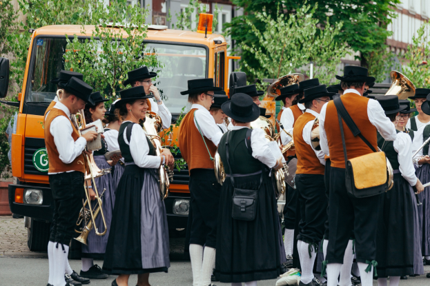 Gruppe von Menschen in traditioneller bayrischer Tracht, die marschieren, Musikinstrumente spielen und Taschen tragen, mit Bäumen und Gebäuden im Hintergrund und einem geparkten Fahrzeug am Straßenrand.