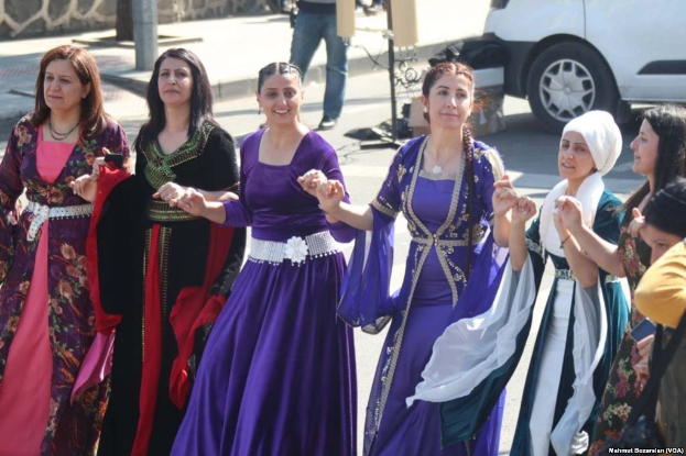 Eine Gruppe von Frauen in bunten Kleidern tanzt auf der Straße, mit einer Person im Hintergrund, die eine Kamera hält, und einem Fahrzeug, einer Wand und Pfosten im Bild.