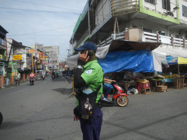 Ein Polizeibeamter in grüner und blauer Uniform und einer blauen Mütze steht an der Straße und spricht in ein Handy, während hinter ihm Fahrzeuge fahren, rechts von ihm Stände mit verschiedenen Gegenständen und im Hintergrund ein Gebäude mit Fenstern und ein bewölkter Himmel zu sehen sind.