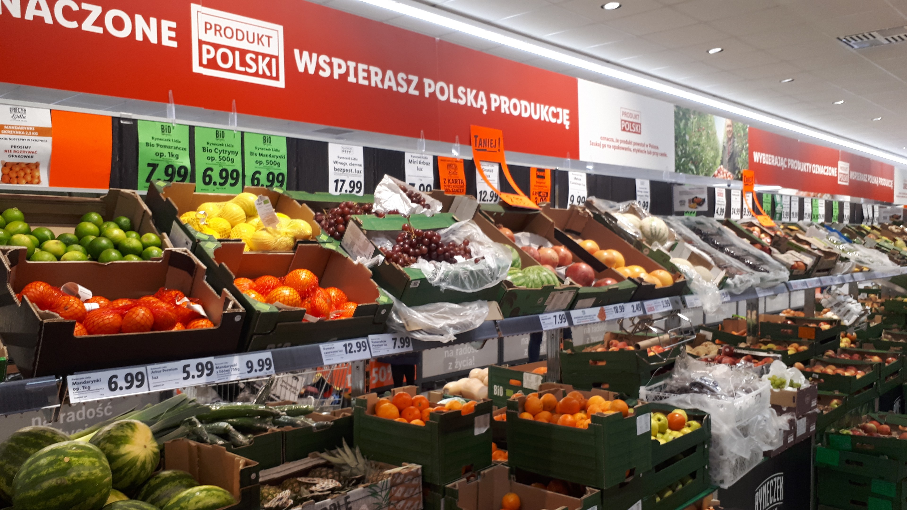 Ladeninneres mit Kartonschachteln verschiedener Früchte und Gemüse, jeweils mit Preisschildern, an der Wand montierten Tafeln mit Text und Bildern sowie Deckenleuchten.