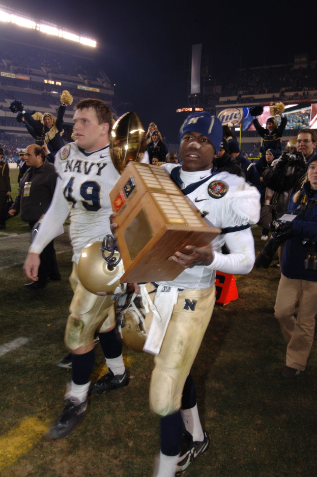 Ein Fußballspieler hält einen Pokal auf einem Feld, umgeben von einer Menge in Sportkleidung, mit Schildern, Lichtern und einem klaren blauen Himmel im Hintergrund.
