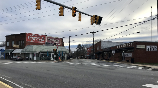 Stadtstraßenkreuzung mit Ampel, Fahrzeugen, Gebäuden mit Namensschildern, Strommasten mit Drähten, Bäumen und einem bewölkten Himmel.