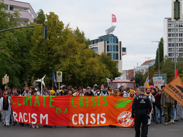 Eine Gruppe von Menschen marschiert eine baumbestandene Straße entlang, die ein Banner mit der Aufschrift "Klima-Krise ist eine Krise" trägt, mit parkenden Fahrzeugen, Schildern, Gebäuden und einem klaren blauen Himmel im Hintergrund.