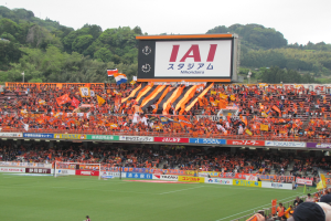 Ein Fußballspiel wird in einem Stadion mit einer großen Menge, grünem Gras, einem Torpfosten, Bannern, Fahnen, einem großen Bildschirm, Bäumen und einem klaren blauen Himmel gespielt.