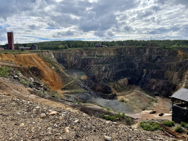 Großer Tagebau inmitten eines Schotterfeldes umgeben von Bäumen, Gebäuden und einem Schuppen, mit bewölktem Himmel und Boden voller Steine und Pflanzen.