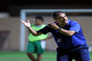 Ein Mann in einem blauen T-Shirt und Hose steht auf einem Fußballfeld und hält ein Mikrofon in der Hand, während er in Richtung der Spieler gestikuliert. Im Hintergrund ist eine Person in einem grünen Kleid und ein Torpfosten zu sehen.