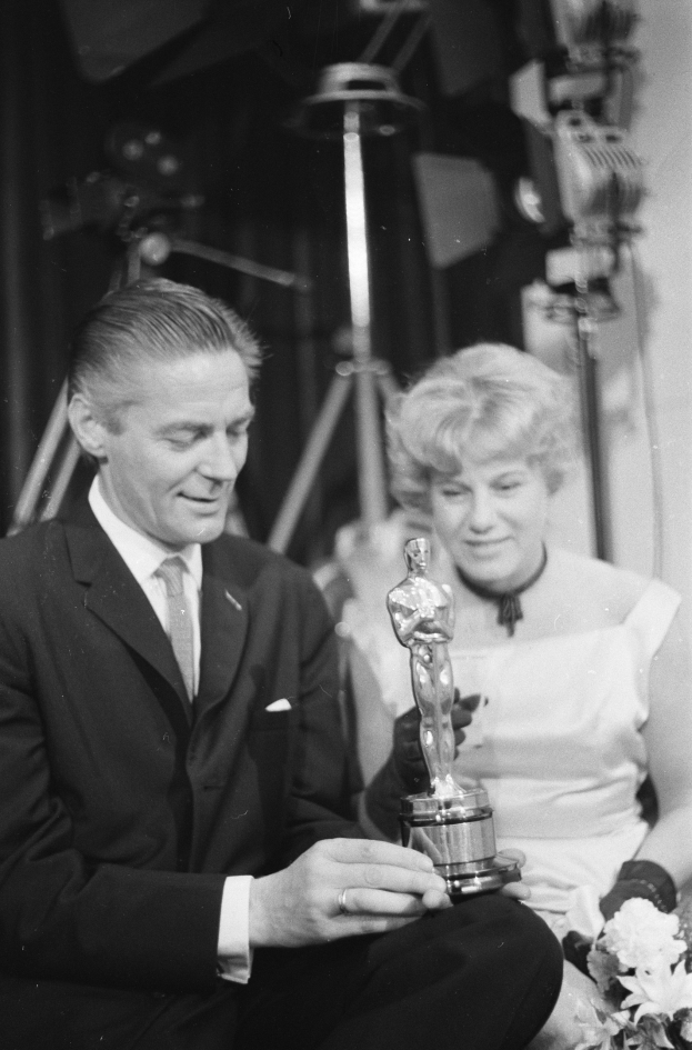 Ein Schwarz-Weiß-Bild eines formell gekleideten Mannes, der eine Oscar-Statue hält und eine Frau, die einen Blumenstrauß hält, nebeneinander sitzen mit beleuchteter Kulisse im Hintergrund.