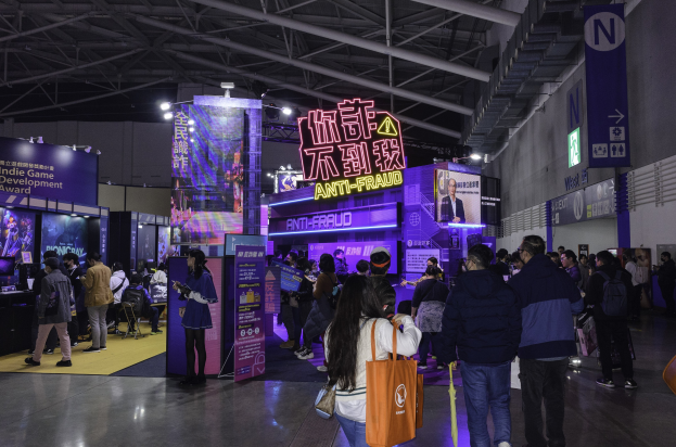 Eine große Gruppe von Menschen steht vor einem Messestand, der mit "Indie Game Development Award 2019" beschriftet ist, wobei einige Taschen und verschiedene Displays, Bildschirme und Tafeln im Hintergrund zu sehen sind.