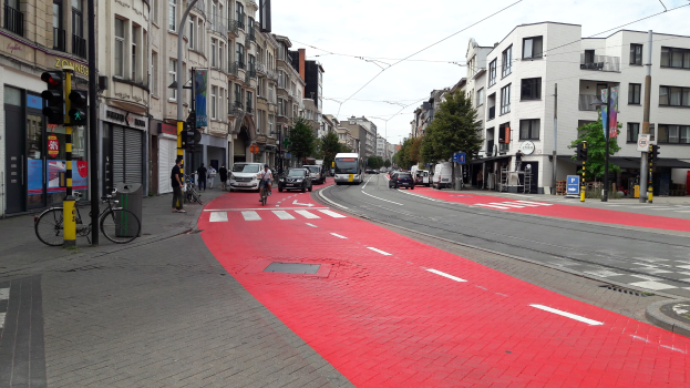 Rote Fahrradspur neben einer Stadtstraße mit Fahrzeugen, Fußgängern, Gebäuden, Bäumen und einem klaren blauen Himmel.
