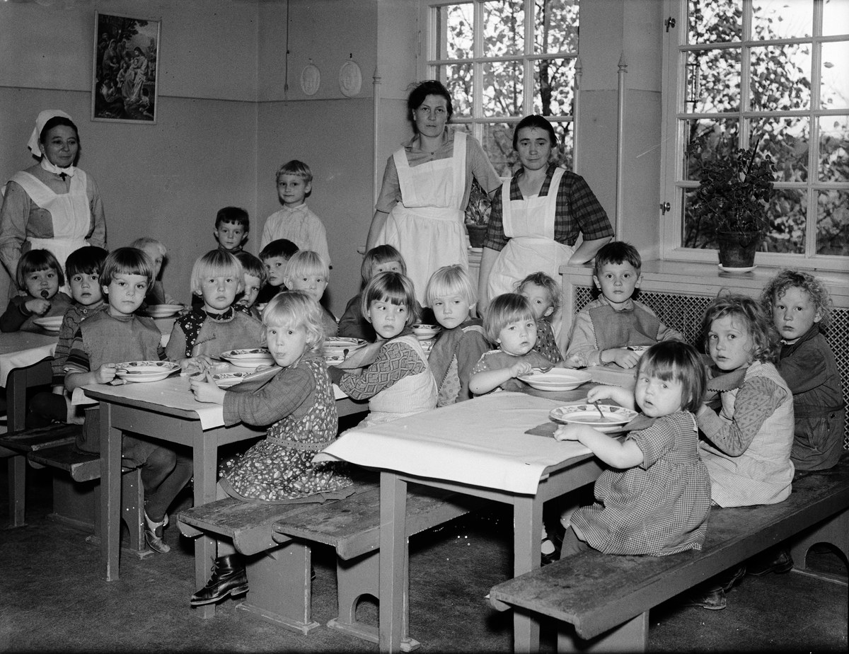 Kinder sitzen an Tischen in einem Klassenzimmer mit Tellern voller Essen, umgeben von Bänken, einem Rahmen an der Wand und Fenstern mit Pflanzen rechts daneben.