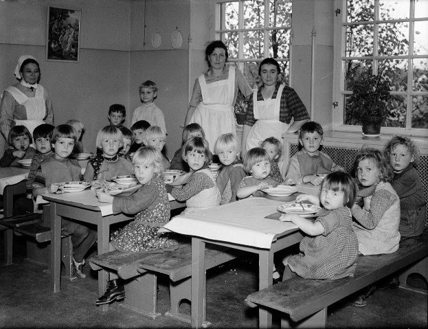 Kinder sitzen an Tischen in einem Klassenzimmer mit Tellern voller Essen, umgeben von Bänken, einem Rahmen an der Wand und Fenstern mit Pflanzen rechts daneben.