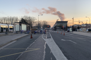 Eine Stadtstraße mit Absperrbaken, einem Bus, Laternen, Bäumen, Gebäuden, Kränen und Rauch, der aus einem entfernten Gebäude aufsteigt, unter einem Himmel.