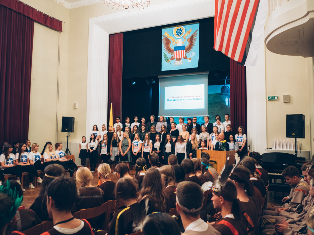Eine Gruppe von Menschen steht vor einem sitzenden Publikum an einem Podium mit Mikrofon, flankiert von Fahnen und Lautsprechern, während einer 2. Woche der Kunst- und Kulturveranstaltung, mit einem Bildschirm, der Text und ein Bild anzeigt, sowie Vorhängen und einem Kronleuchter im Hintergrund.