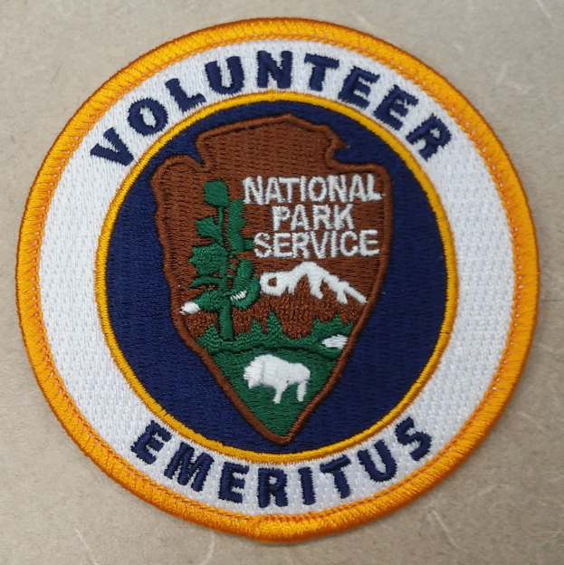 Runder Aufnäher mit blauem Rand und weißem Hintergrund, der den schwarzen Schriftzug "Volunteer National Park Service Emeritus" in der Mitte eines gelben Kreises zeigt.