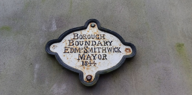 Metallplakette an der Seite eines Gebäudes, die die Borough-Grenze markiert, mit "Edm Smithwick Mayor" in fetten weißen Buchstaben auf schwarzem Hintergrund.