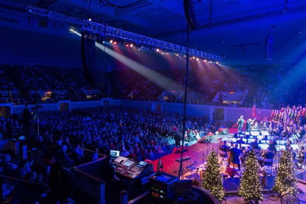 Eine große Menschenmenge in einem geschmückten Auditorium schaut einer Band auf der Bühne zu, die von Weihnachtsbäumen, Lautsprechern und festlicher Beleuchtung umgeben ist.