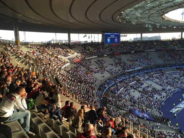 Große Menschenmenge auf Tribüne bei einem Fußballspiel, mit Bühne rechts, Fahnen, Masten, einem Bildschirm und der Allianz Arena in München, Deutschland im Hintergrund.