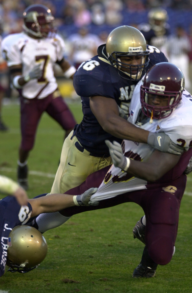 Zwei Fußballer mit Helmen und Uniformen stoßen auf dem Feld zusammen, während andere zuschauen.