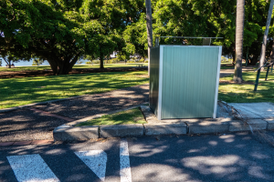 Transportable Toilette am Straßenrand in einem Park, umgeben von Bäumen und Gras, mit einem Gewässer und einem klaren blauen Himmel im Hintergrund.