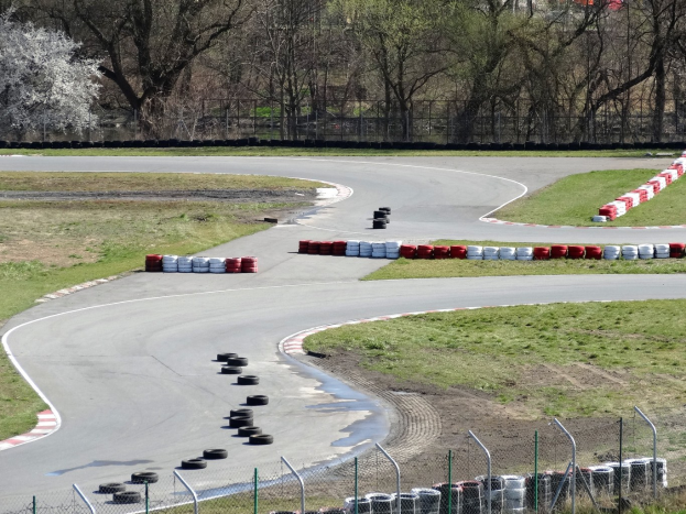 Rennstrecke mit zahlreichen Reifen entlang ihrer Ränder, umgeben von Gras und Maschendrahtzaun, mit Bäumen im Hintergrund.