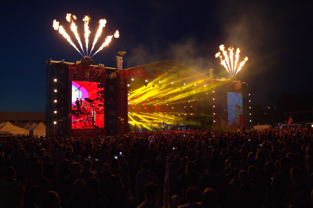 Eine große Menschenmenge auf einem Musikfestival mit einer hell erleuchteten Bühne mit Musikern und Bannern, Zelte links und ein Feuerwerk, das den dunklen Himmel erhellt.