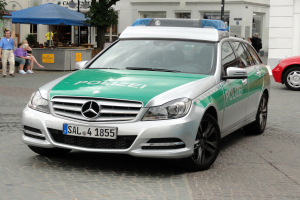 Ein Polizeiauto, genauer ein Mercedes-Benz C-Klasse W204 mit glänzender schwarzer Lackierung, steht auf der Straße neben einem roten Auto, mit verschiedenen Gebäuden, Bäumen und Menschen im Hintergrund.