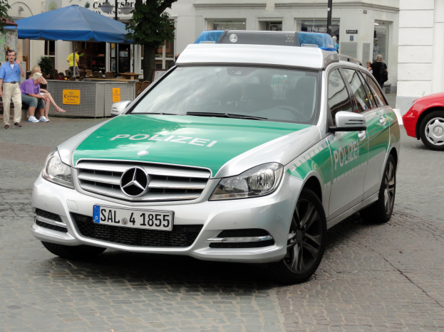 Ein Polizeiauto, genauer ein Mercedes-Benz C-Klasse W204 mit glänzender schwarzer Lackierung, steht auf der Straße neben einem roten Auto, mit verschiedenen Gebäuden, Bäumen und Menschen im Hintergrund.