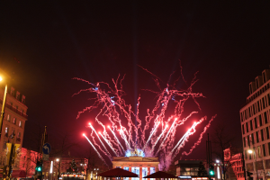 Eine belebte Stadtstraße in Berlin am Neujahrstag, voller Menschen, Fahrzeuge und festlicher Dekorationen, mit Feuerwerk, das den Nachthimmel und Gebäude erhellt.