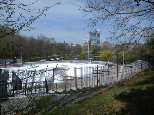 Eisbahn in einem Park umgeben von einem Metallzaun, Gras, Bäumen, Fahrzeugen, Straßenlaternen, einem Gewässer, Gebäuden und einer bewölkten Himmel.