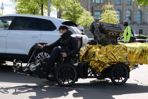 Ein Mann in einer schwarzen Jacke und Mütze sitzt in einem Rollstuhl mit einem großen Motor am Rücken, umgeben von Fahrzeugen auf einer Straße mit Bäumen, Gebäuden und Pfählen im Hintergrund unter einem klaren blauen Himmel.