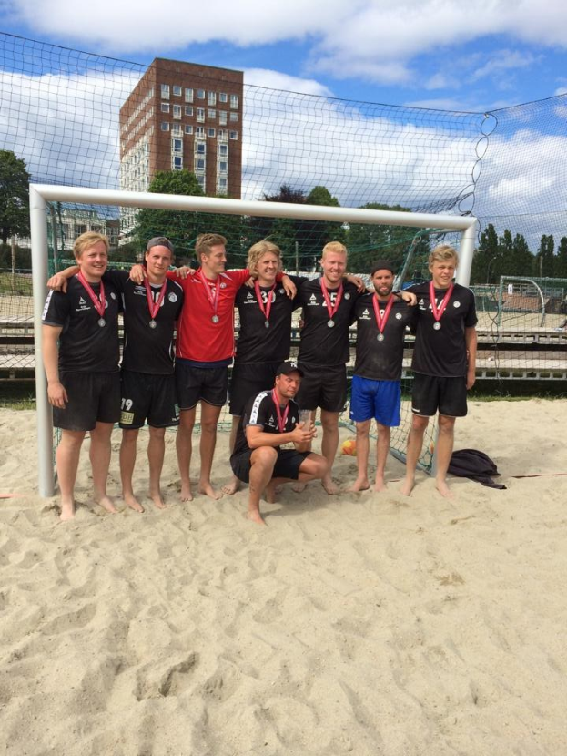 Mannergruppe in Sportbekleidung auf einem sandigen Strand, mit Medaillen und umgeben von einem Netz, mit Bäumen, Gebäuden und einem bewölkten Himmel im Hintergrund.