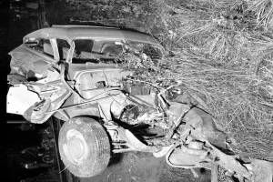 Ein Schwarz-Weiß-Foto eines schwer beschädigten Autos am Straßenrand, mit eingedellter Front, zersplitterten Fenstern, einem Baum im Hintergrund und verstreuten Gegenständen rund um die Szene.