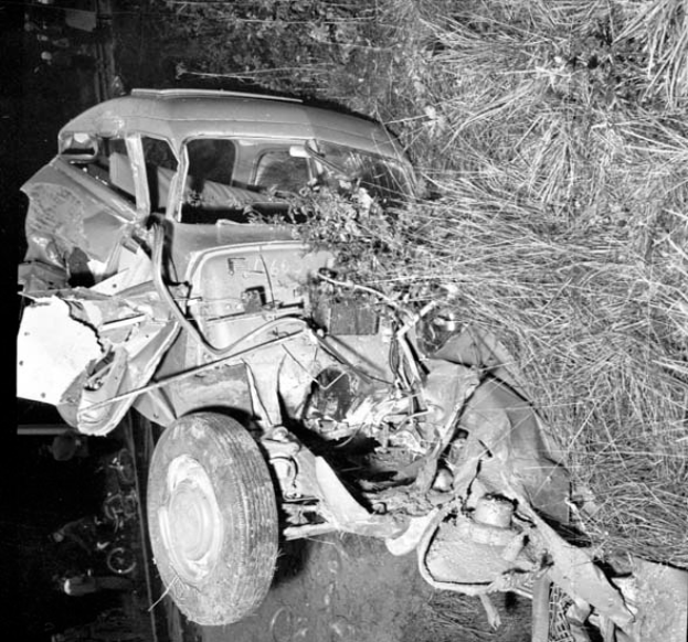 Ein Schwarz-Weiß-Foto eines schwer beschädigten Autos am Straßenrand, mit eingedellter Front, zersplitterten Fenstern, einem Baum im Hintergrund und verstreuten Gegenständen rund um die Szene.