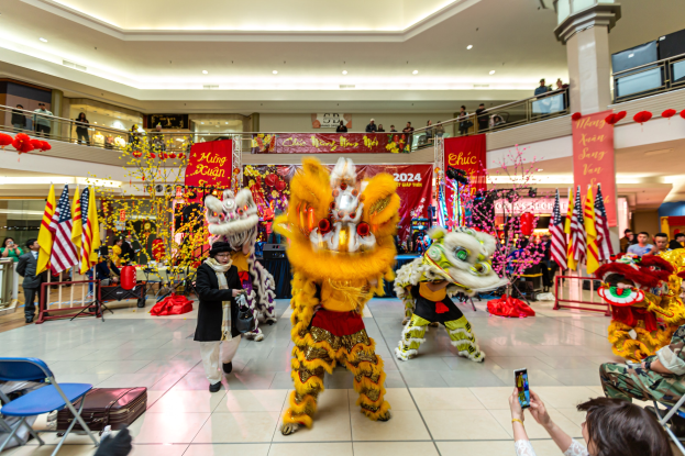 Lebendiges chinesisches Neujahrsfest in einem Einkaufszentrum mit tanzenden Menschen in Kostümen, anderen auf Stühlen sitzend und Mobiltelefone haltend sowie verschiedenen Dekorationen, Flaggen und Bannern im Hintergrund.